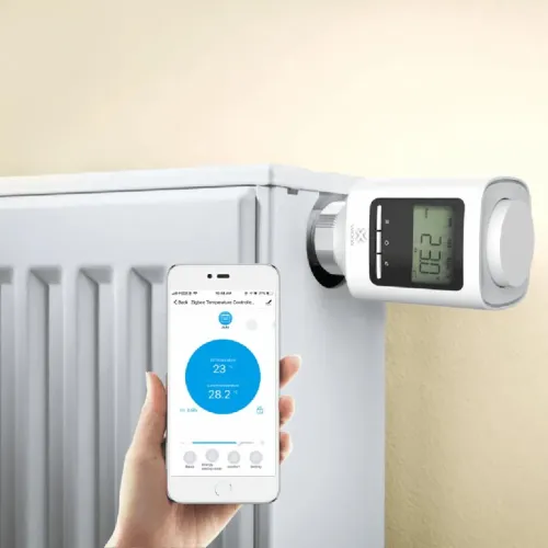 WOOX smart radiatorventil med sidemonteret display installeret på en hvid radiator, ved siden af en håndholdt mobil med app.